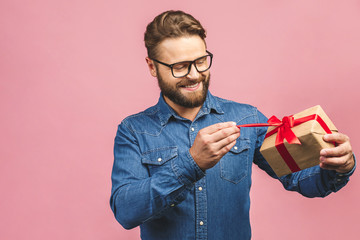 Happy holiday, my congredulations! Portrait of an attractive casual man giving present box and looking at camera isolated over pink background.