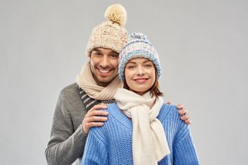 people, christmas and winter clothes concept - happy couple in knitted hats and scarves over grey background