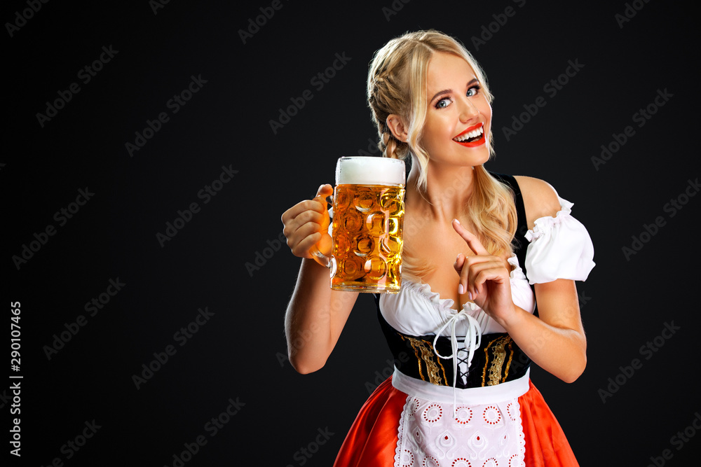 Wall mural Smiling young sexy oktoberfest girl waitress, wearing a traditional Bavarian or german dirndl, serving big beer mug with drink isolated on black background.