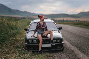 Rear view of young girl in gray short denim shorts is repairing the car. In shorts near a black car with open hood. Problems with the car in the road trip. brunette repairing the engine