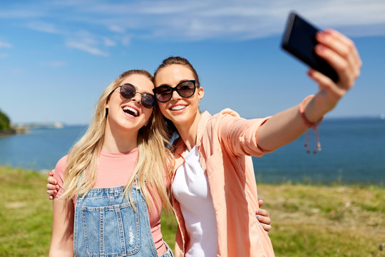 leisure and friendship concept - happy smiling teenage girls or best friends in sunglasses hugging and taking selfie by smartphone at seaside in summer