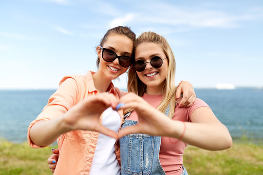 leisure and friendship concept - happy smiling teenage girls or best friends in sunglasses at seaside in summer hugging and making hand heart gesture