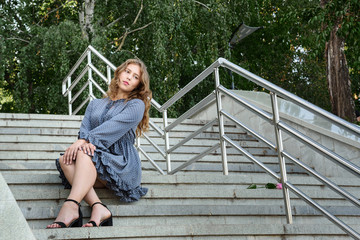 Brunette girl sitting on stairs in the city. Girl in blue dress with long hair on the street