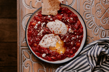 Morning breakfast. Tomato shakshuka with two poached eggs on a plate. Crispy sourdough bread on the side. Coffee in french press.