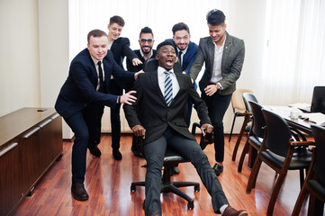 Six multiracial business mans standing at office and roll man on chair. Diverse group of male employees in formal wear having fun.