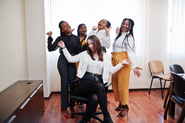 Five multiracial business womans standing at office and roll woman on chair. Diverse group of female employees in formal wear having fun.