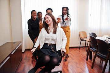 Five multiracial business womans standing at office and roll woman on chair. Diverse group of female employees in formal wear having fun.