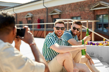 leisure, male friendship and people concept - man with camera photographing his friends drinking...