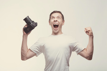 Fototapeta na wymiar Emotional Man with camera isolated on light background. Young man holding digital camera with victory screaming. Lifestyle, travel and technology concept. Glad boy in white t-shirt with terrific face