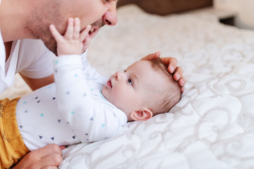 Close up of cute six months old baby boy lying on bed and playing with his dad. Dad talking to son...