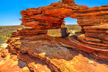 Blonde caucasian woman sitting inside the iconic rock arch in red sandstone of Nature's Window, Kalbarri National Park. Caucasian young girl in Western Australia looking australian outback landscape.