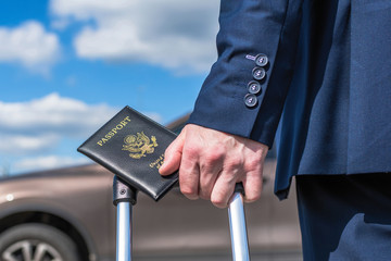 Man in a blue suit with a suitcase and american passport at the airport parking on the backdrop of his car. Business trip concept