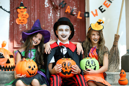 Two Young Girls And Boy In Halloween Costumes Sitting On Porch And Holding Pumpkin Buckets Full Of Candies