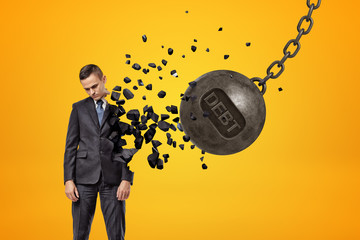 Black wrecking ball with word DEBT on it hitting sad young businessman and breaking half of his...