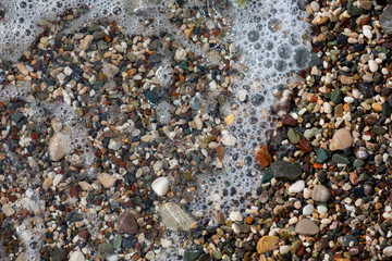 Thin white foam and the many small bubbles on the glittering sea water with the colorful pebbles underneath