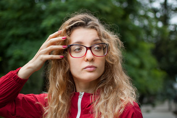 Young beautiful woman with blonde curly hair in dark red hipster jacket and eyeglasses posing...