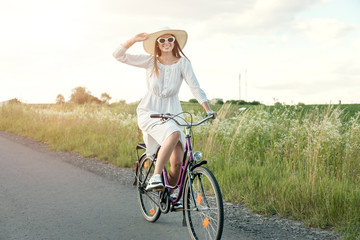 Attractive cyclist girl in white dress and straw hat posing in the sunset