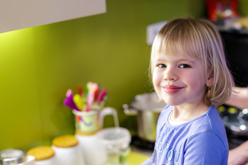 Obraz na płótnie Canvas Pretty happy little smiling girl with blond hair in purple shirt standing in kitchen.