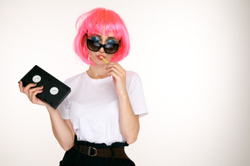 Retro girl in the glasses and pink wig holding black cassette on the white background