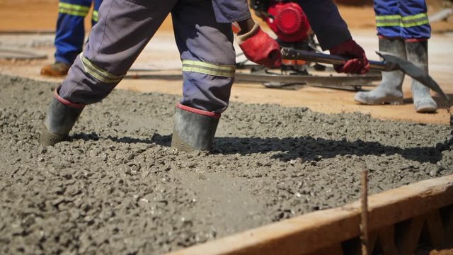 Medium slow motion shot of a rubber boot-wearing construction worker spreading out wet cement with shovel during afternoon.