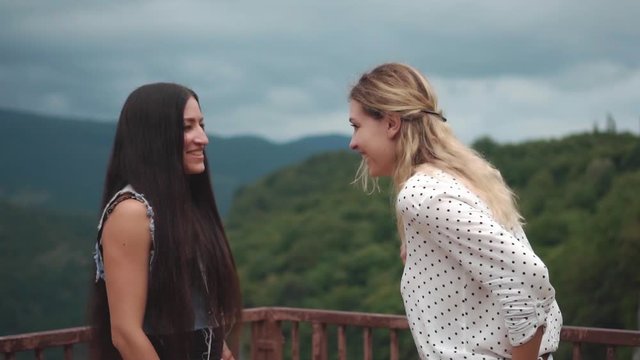 Two women in an informal setting are discussing business, amid a mountain landscape, amiably clapping hand in hand. The gesture is agreed. Blonde and brunette in casual clothes are standing on bridge