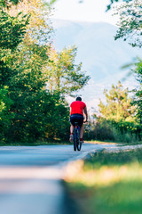 Fototapeta premium Fit cyclist rides his bicycle (bike) on an empty road in nature wearing a baseball hat and red t-shirt.