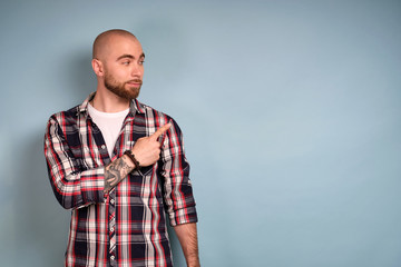 A bearded guy in a plaid shirt stands on a blue background and looks quite and shows his index fingers to the side