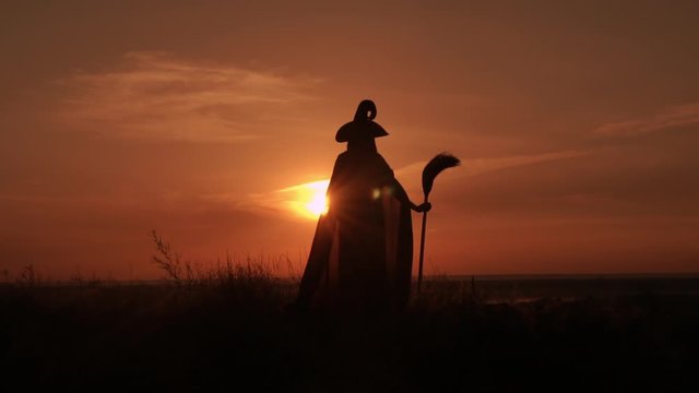 woman in halloween costume sorceress holding besom posing on the hill sky with yellow sun above the city slow motion