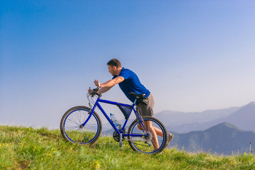 Fit male mountain biker pushing his bike uphill while looking exhausted on a sunny summer day.
