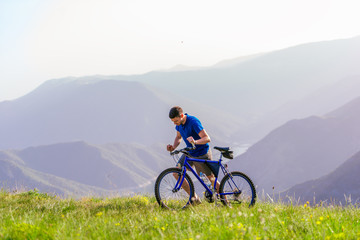 Fototapeta premium Tired fit mountain biker pushing his bike uphill at the top of the mountain on a sunny day with amazing view on a blue river.