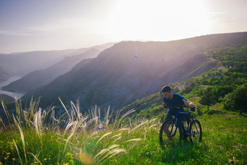 Obraz premium Perspective of a fit mountain biker pushing his bike uphill with amazing view on a forest, river and mountains in the background. Amazing green nature at sunset.