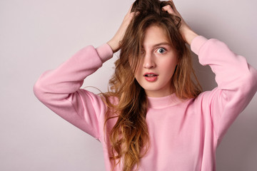 A blonde girl in a pink sweatshirt stands on a white background and looks in horror at the frame ruffling her hair