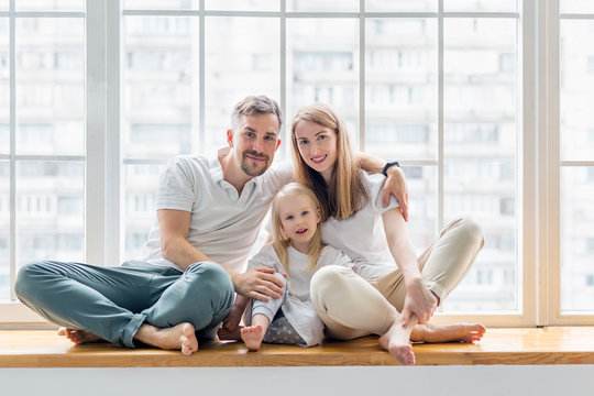 Happy Family Sitting On Windowsill Together. Family Smiling While Sitting In Front Of Window