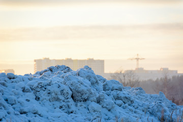 Winter landscape. A mountain of snow against the backdrop of a sunset, cityscape and tower crane.