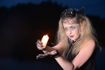 A woman with tousled hair holds a burning candle in her hands. Halloween makeup witch. Night photo of a girl with a candle.