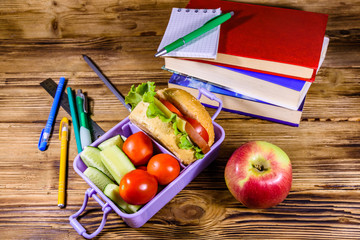 Notepad, pens, ripe apple, stack of books and lunch box with hamburger, cucumbers and tomatoes on wooden table