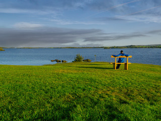 Man sitting on a wooden bench at lake Corrib, looking at dragon throwing flame looking cloud on horizon. sunny warm day.
