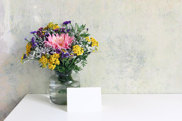 Glass vase with beautiful colorful bouquet of dahlia, tansies and aster flowers. Grunge old wall background. Feminine wedding, birthday mock-up scene with blank greeting card. Empty copy space.