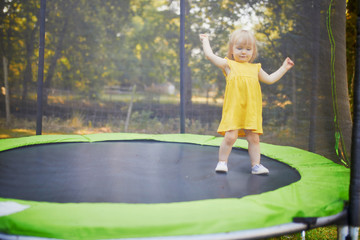 Adorable toddler girl in yellow dress having fun on a trampoline on a summer day