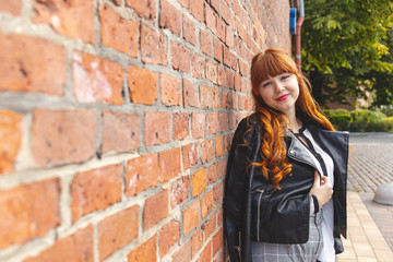 Cute young woman in a black leather jacket with curly red hair and bangs smiles against a brick wall