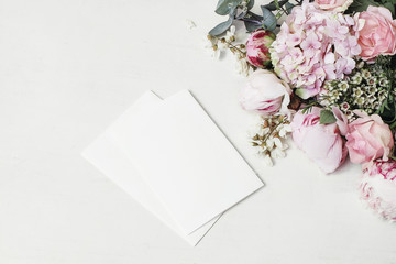 Feminine wedding, birthday mock-up scene. Decorative floral corner of peony, hydrangea, roses and locust flowers. Blank paper greeting cards on white wooden table background. Fat lay, top view