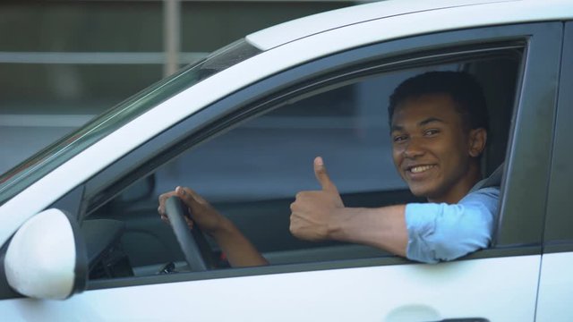 Mixed-race teenager sitting on driver seat of car showing thumbs-up, driving