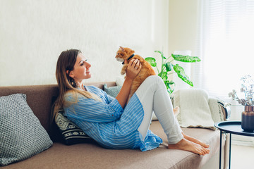 Playing with cat at home. Young woman sitting on couch and hugging pet.