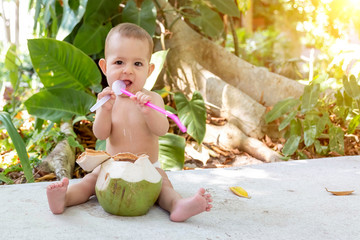 Teething and salivation infant baby at tropical vacation. Eats and drinks green young coconut. Sits on a ground and gnaw a spoon. Jungles on background.