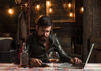 Drinking man is cheking his phone. Man with beard holds glass brandy. Man holding a glass of whisky. Sipping whiskey. Degustation, tasting. Macho is drinking whiskey by his laptop. Tired.