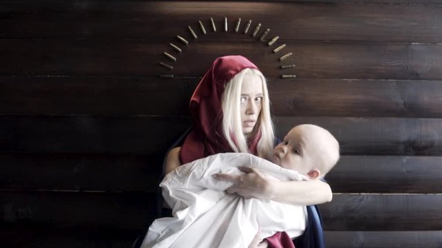 Close up of a scared young woman weaing red hood holding a baby in white sheet on wooden wall background. Footage. An image of Virgin Mary and Jesus, religion concept.