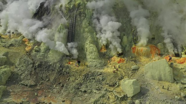 Cinematic aerial view of sulfur miners mining sulfur while it spews toxic sulfur gas clouds at the crater of Kawah Ijen volcano, East Java, Indonesia.