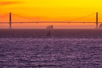 The sun sets over the Golden Gate Bridge as a sailboat and a large container ship set off on the horizon. 