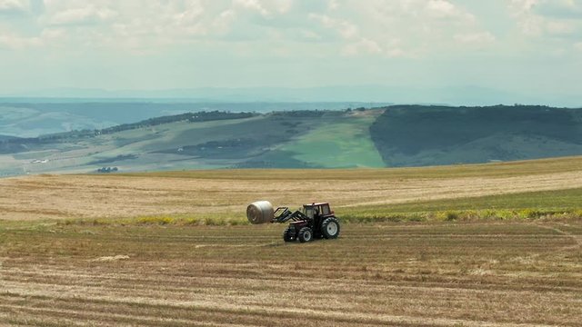 Aerial shot of a hay tractor collecting and loading hay bales in a beautiful Italian landscape with wind turbines in the background  
