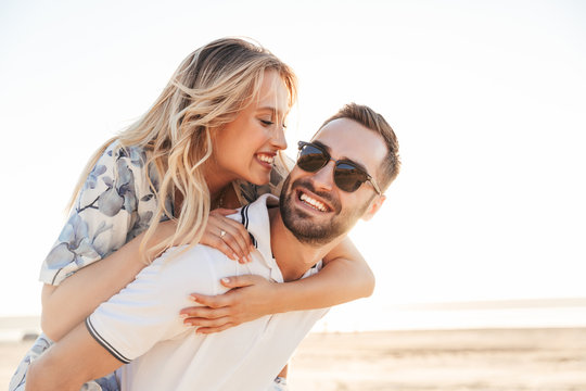 Photo of unshaven happy man smiling and giving piggyback ride seductive woman while walking on sunny beach